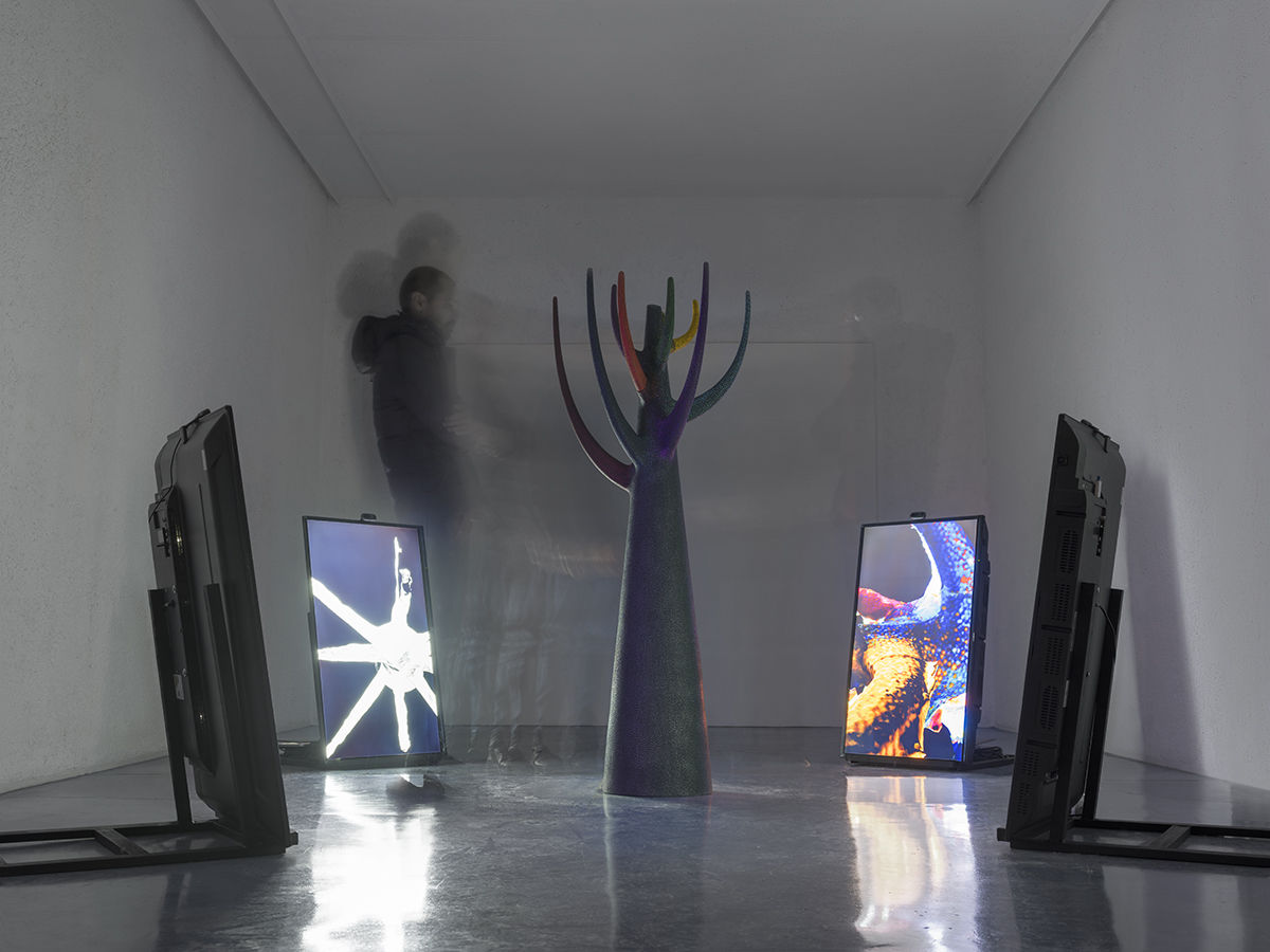 vue de l'installation, sculpture au centre du salle noire, elle représente une sorte d'arbre géométrique, autour quatre écrans projetent des images 3D de la sculpture et réagissent à la présence du public, une personne est à côté de la scultpure