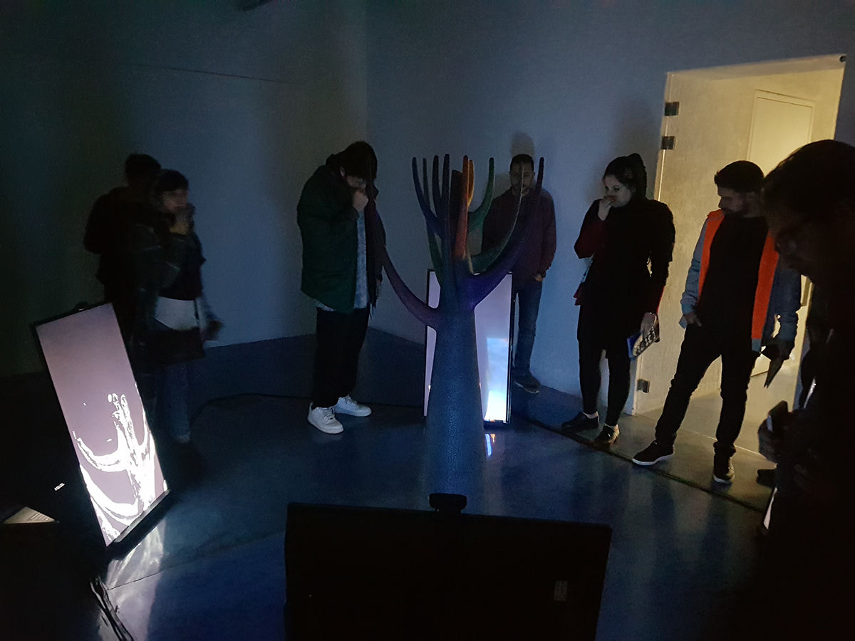 vue de l'installation, sculpture au centre du salle noire, elle représente une sorte d'arbre géométrique, autour quatre écrans projetent des images 3D de la sculpture et réagissent à la présence du public, plusieurs personnes entourent le dispositif